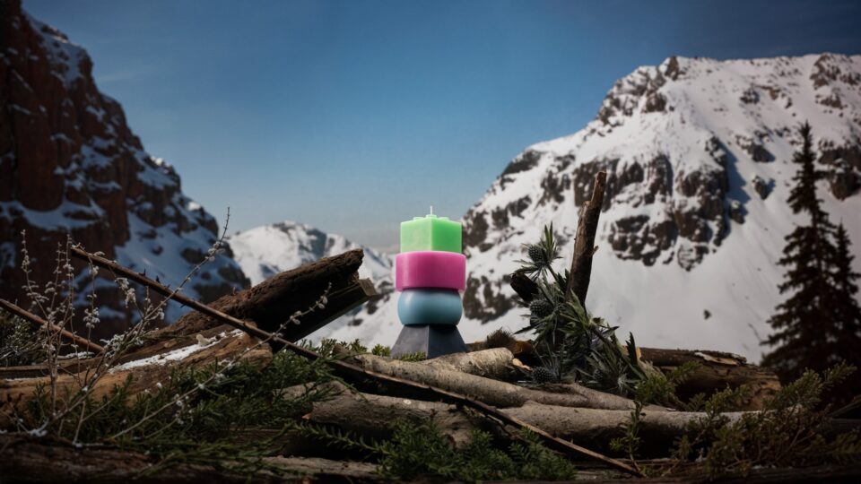 Colorful stacked candle standing on a log with a snow-covered mountain and clear sky in the background creating a scenic and peaceful outdoor winter setting.