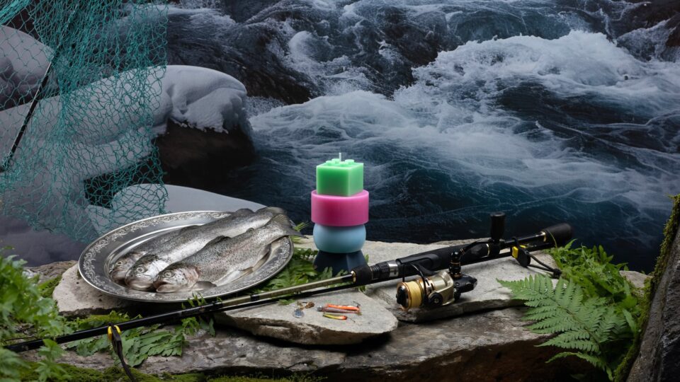 Fishing scene by a river with a rod, reel, and caught fish on a plate. Vibrant candles and ferns add color to the serene setting. Perfect for nature and angling enthusiasts.